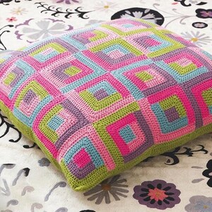 Könnte beinhalten: Ein quadratisches Häkelkissen mit einem geometrischen Muster. Das Design zeigt konzentrische Quadrate in Rosa-, Lila-, Blau- und Grüntönen mit einem grünen Rand. Das Kissen liegt auf einer Oberfläche mit floralem Muster.