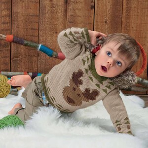 May include: A tan knitted baby sweater with a brown reindeer design on the front. The sweater has green accents and a faux fur collar. The baby is wearing khaki pants and is lying on a white fluffy surface.
