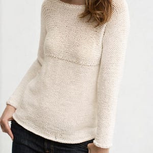 Könnte beinhalten: Cremefarbener Strickpullover mit rundem Ausschnitt und langen Ärmeln. Der Pullover hat ein strukturiertes Strickmuster und ein horizontales Banddetail über der Brust. Das Kleidungsstück besteht aus einem weichen, leichten Material.