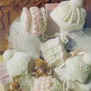 Puede incluir: Una colección de gorros y capotas de bebé de ganchillo hechos a mano, en color blanco roto y rosa. Algunos gorros tienen un pompón en la parte superior. También hay un pequeño osito de peluche y un conejito.