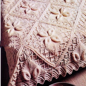 May include: A cream-colored, knitted blanket with a floral pattern. The blanket features raised, three-dimensional flower designs and a decorative, scalloped edge. The intricate knitwork creates a textured surface, perfect for a bedspread or decorative throw.