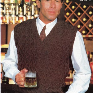 Puede incluir: Un chaleco marrón de punto trenzado con cuello en V, usado sobre una camisa blanca con cuello y una corbata estampada. La imagen incluye texto que indica medidas de 86-112 cm. La persona sostiene un vaso de cerveza.