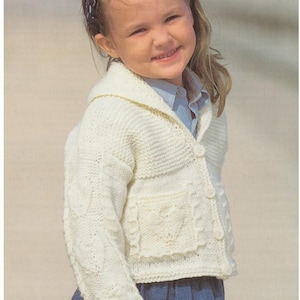 May include: A cream-colored, knitted cardigan with a shawl collar and heart-shaped pocket detail. The cardigan has button closures and cable knit sleeves. The child is wearing a blue headband and a blue shirt.