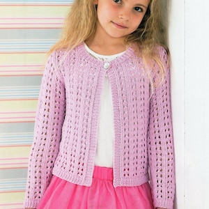 Peut inclure: Un cardigan lilas clair avec un motif ajouré et des manches longues. Le gilet en tricot a une fermeture à un seul bouton et est porté avec une jupe rose. Le cardigan est en tissu tricoté doux.