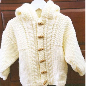 Puede incluir: Chaqueta con capucha de punto color crema con cierres de palanca de madera. La chaqueta presenta un diseño de punto trenzado en la parte delantera y un patrón de punto texturizado. La chaqueta se muestra en una percha blanca.