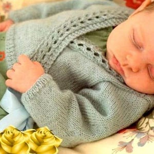 May include: A sleeping baby wrapped in a light blue knitted sweater. The sweater has a delicate openwork design along the neckline and cuffs. The baby is resting on a floral patterned blanket, with a green floral fabric visible.