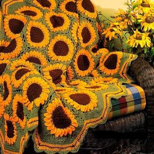 Op de afbeelding: Een handgemaakte gehaakte deken met een zonnebloemontwerp. De deken heeft een groene achtergrond met bruine en gele zonnebloemen. De deken is gedrapeerd over een rieten stoel met een geruit kussen.