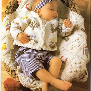 May include: A sleeping baby in a wicker basket, wearing a white cardigan with embroidered designs and checkered shorts. The baby is resting on a knitted blanket with a matching pattern. A gingham bow headband completes the outfit.