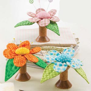 Op de afbeelding: Drie kleurrijke bloemvormige speldenkussens. De bloemen hebben stoffen bloemblaadjes in oranje, roze en blauw, met groene bladeren. Elke bloem staat op een houten spoel. Spelden zijn in het midden van elke bloem gestoken.