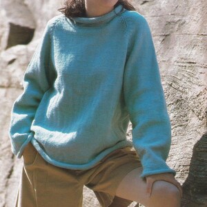 May include: A light blue knitted jumper with a high neckline and long sleeves. The jumper is worn with tan shorts. The model is standing in front of a rock wall.
