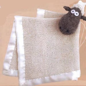 May include: A cream-colored knitted baby blanket with a white satin border. A small, brown crocheted sheep with white accents and large, round eyes rests on the blanket. The blanket is folded, showcasing its texture and design.