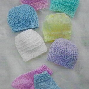 May include: A collection of six hand-knitted beanies in pastel shades of pink, turquoise, blue, yellow, white, and lavender, accompanied by two pairs of matching hand-knitted baby socks in pink and blue, all featuring a textured knit pattern.