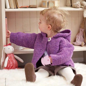 May include: A young child wearing a purple hooded knit sweater with a single white button. The child is reaching for a book on a shelf. A stuffed bunny in a pink dress and a toy are in the foreground. The scene is set in a cozy, well-lit room.