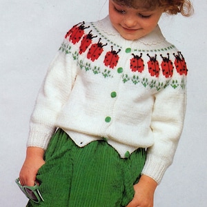 Op de afbeelding: Een witte gebreide cardigan met rode lieveheersbeestjesmotieven en groene knopen. De cardigan wordt gecombineerd met een groene corduroy rok. Het lieveheersbeestjespatroon herhaalt zich op de borst en mouwen.