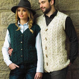 May include: A man and woman wearing knitted cable-stitch vests. The woman's vest is dark green, the man's is cream. Both vests have wooden buttons. They stand against a stone wall, the woman wearing a hat.