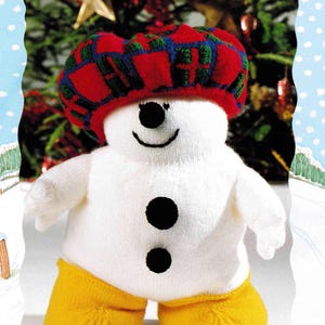 May include: A knitted snowman wearing a red, green, and blue plaid hat and yellow shorts. The snowman has a black button nose and two black buttons on its white body. A smiling face completes the design.