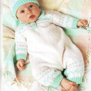 May include: A baby in a knitted white romper with mint green and pink detailing, and a matching hat. The romper has long sleeves and legs. The baby is lying on a soft, patterned blanket.