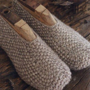 Op de afbeelding: Een paar handgemaakte, lichtbruine gebreide pantoffels met houten schoenvormen erin. De pantoffels zijn gemaakt van dik, getextureerd garen en liggen op een verweerd houten oppervlak. De stijl is gezellig en rustiek.