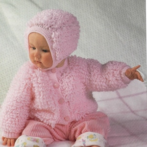 Peut inclure: Un bébé vêtu d'un ensemble tricoté rose clair, comprenant un bonnet, un cardigan et un pantalon. Le cardigan se ferme avec des boutons, et le pantalon a des poignets à motifs. La texture tricotée est duveteuse.