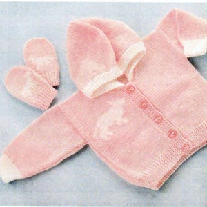 Puede incluir: Un conjunto de bebé de punto rosa claro que incluye una rebeca con capucha con botones, manoplas a juego y una capucha. El conjunto presenta ribetes blancos y un diseño blanco en la rebeca.