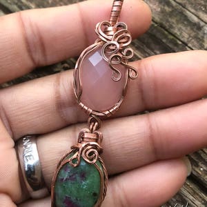 May include: A handcrafted copper pendant featuring two gemstone elements. The top stone is a faceted pink oval, and the bottom is a green and red oval. The pendant is wire-wrapped with intricate copper detailing. The pendant is held in a hand.