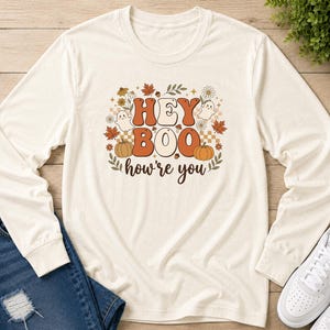 May include: A cream-colored long-sleeve shirt featuring a graphic print with the text "HEY BOO how're you" in brown and orange. The design includes illustrations of two small ghosts, two orange pumpkins, autumn leaves, and small flowers.