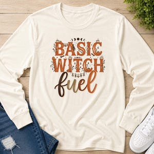 May include: A cream-colored long-sleeve shirt featuring the text BASIC WITCH fuel in brown and orange gradient lettering. The design includes small illustrations of coffee cups, pumpkins, moon phases, and foliage surrounding the bold typography.