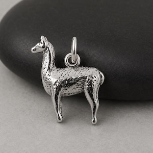 May include: A silver-toned metal charm shaped like a standing llama, featuring a textured surface and a small circular jump ring attached to the top for hanging. The charm has a metallic finish with dark recessed areas that highlight the animal's form.