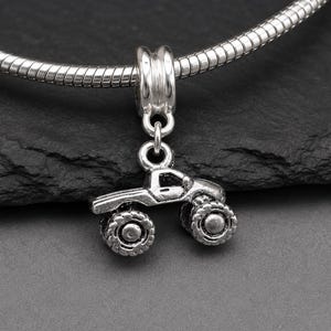 May include: A silver-toned metal charm shaped like a monster truck with large, textured tires and a detailed truck body. The charm is suspended from a silver-toned snake chain bracelet, featuring a cylindrical bail connector at the top.