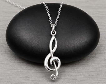 May include: A silver-toned necklace featuring a treble clef pendant suspended from a delicate chain. The pendant has a polished, reflective metallic finish and a smooth, curved design that mimics the shape of a musical clef symbol.