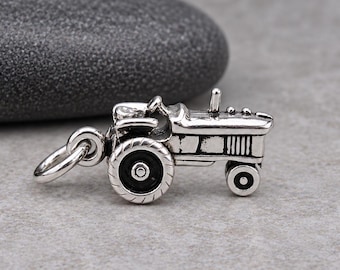 May include: A silver-toned metal charm shaped like a farm tractor, featuring a detailed engine block, a steering wheel, and a large rear wheel with a black centre. The charm includes a small circular jump ring attached to the front for hanging.