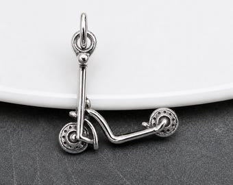 May include: A silver-toned metal charm shaped like a kick scooter, featuring a vertical handlebar stem, a horizontal deck, and two small wheels with detailed spokes. The charm includes a circular jump ring at the top for attachment purposes.
