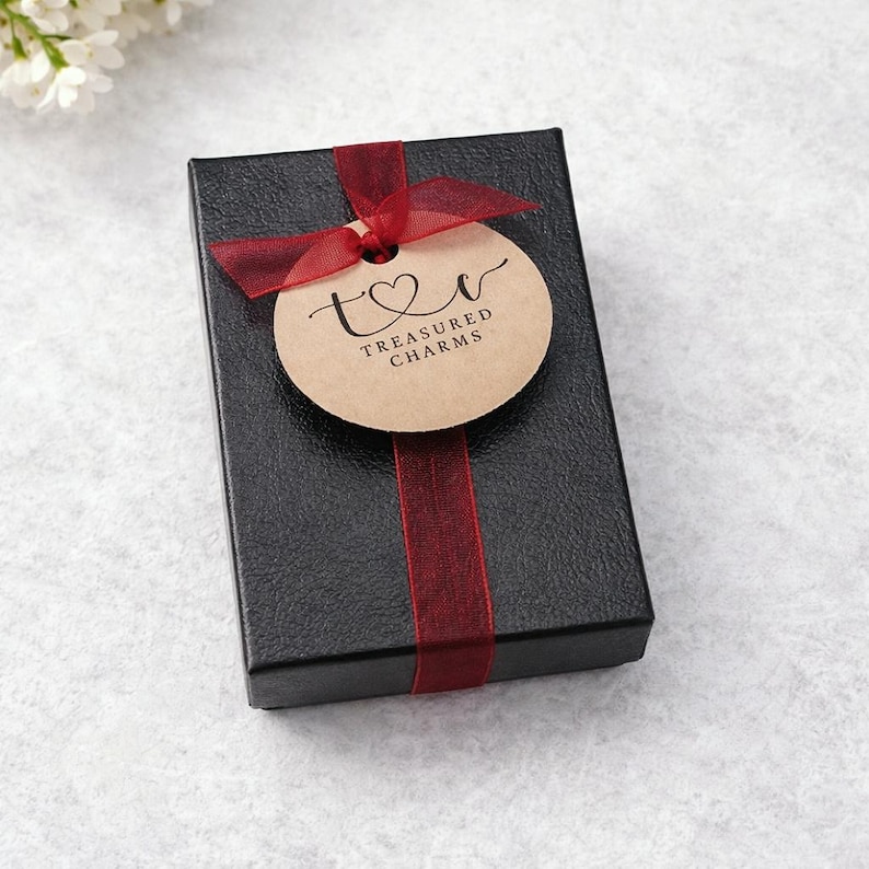 A black gift box with a red ribbon and a round tag that says "Treasured Charms" is placed on a white surface.