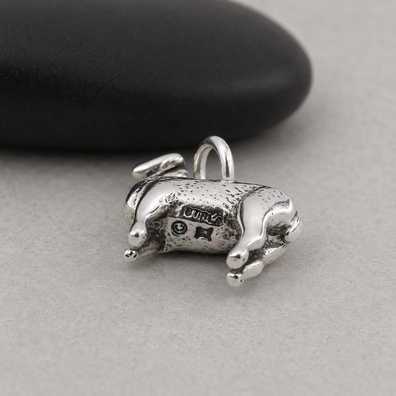May include: A silver-toned metal charm shaped like a small animal, featuring a textured surface and a jump ring at the top. The side of the charm displays the stamped text "CURA" along with a small circular symbol and a four-pointed star mark.