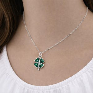 Puede incluir: Collar de plata con un colgante de tr&eacute;bol de cuatro hojas. El tr&eacute;bol es verde con un contorno plateado y est&aacute; suspendido de una delicada cadena de plata. El collar se lleva sobre una camiseta blanca.