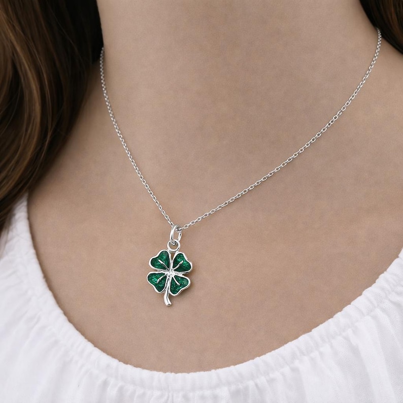 Puede incluir: Collar de plata con un colgante de tr&eacute;bol de cuatro hojas. El tr&eacute;bol es verde con un contorno plateado y est&aacute; suspendido de una delicada cadena de plata. El collar se lleva sobre una camiseta blanca.