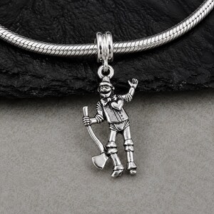 Könnte beinhalten: Ein silberfarbener Metallanhänger in Form einer Figur mit Trichterhut, Fliege und einem Herz auf der Brust, die eine Axt hält. Der Anhänger ist an einem silberfarbenen Schlangenkettenarmband befestigt und weist detaillierte Gravuren auf.