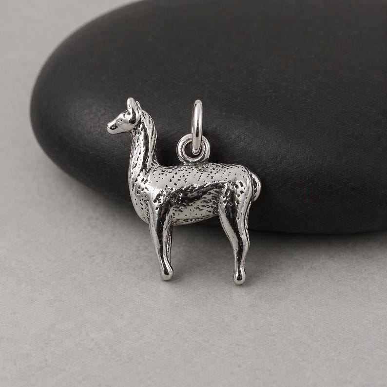 May include: A silver-toned metal charm shaped like a standing llama, featuring a textured surface and a small circular jump ring attached to the top for hanging. The charm has a metallic finish with dark recessed areas that highlight the animal's form.