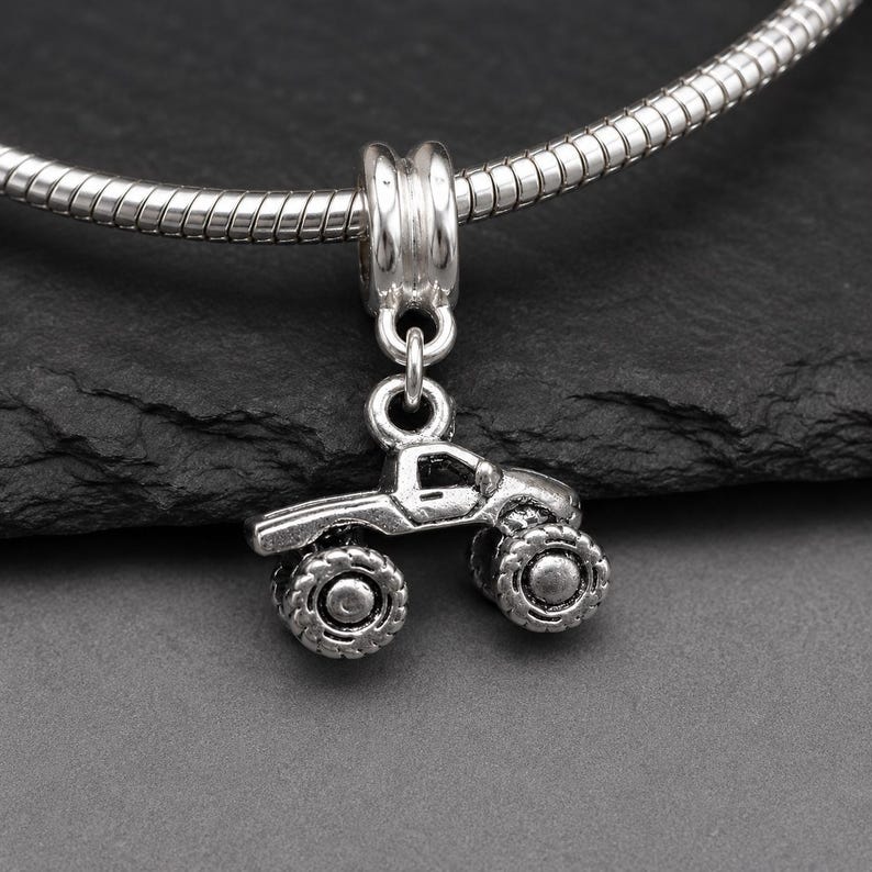 May include: A silver-toned metal charm shaped like a monster truck with large, textured tires and a detailed truck body. The charm is suspended from a silver-toned snake chain bracelet, featuring a cylindrical bail connector at the top.