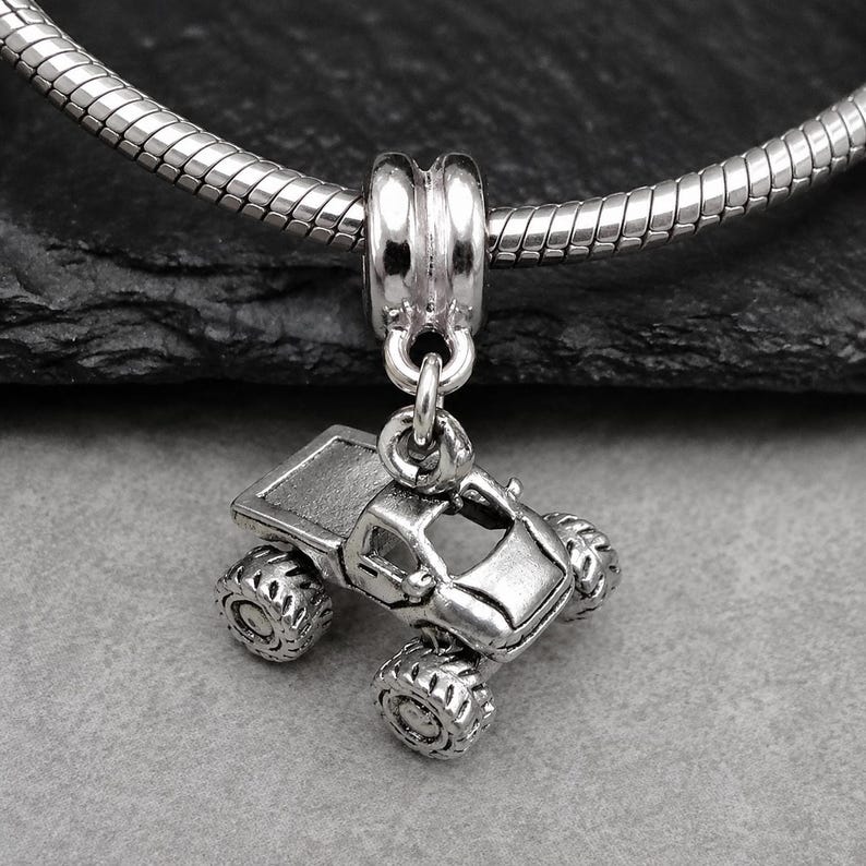 May include: A silver-toned metal charm shaped like a monster truck with large, textured tires and an open-cab design. The charm is suspended from a polished silver-toned metal bail, which is threaded onto a flexible, snake-style silver chain necklace.