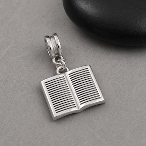 Può includere: Un ciondolo d'argento a forma di libro aperto. Il ciondolo presenta linee dettagliate che rappresentano le pagine e un anello per il fissaggio a un braccialetto o una collana. Il ciondolo è realizzato in metallo lucido e riflettente.