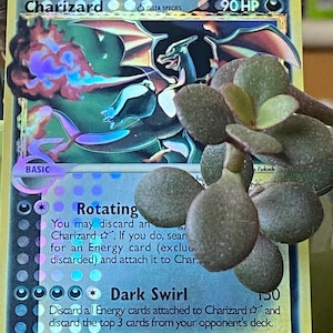 May include: A holographic Charizard Pokemon card from the Delta Species set. The card is yellow and blue with a picture of Charizard breathing fire. The card's name, "Charizard", is printed in black at the top of the card. The card's HP is 90. The card's attacks are "Rotating" and "Dark Swirl". The card's retreat cost is three stars. The card's number is 100/101.