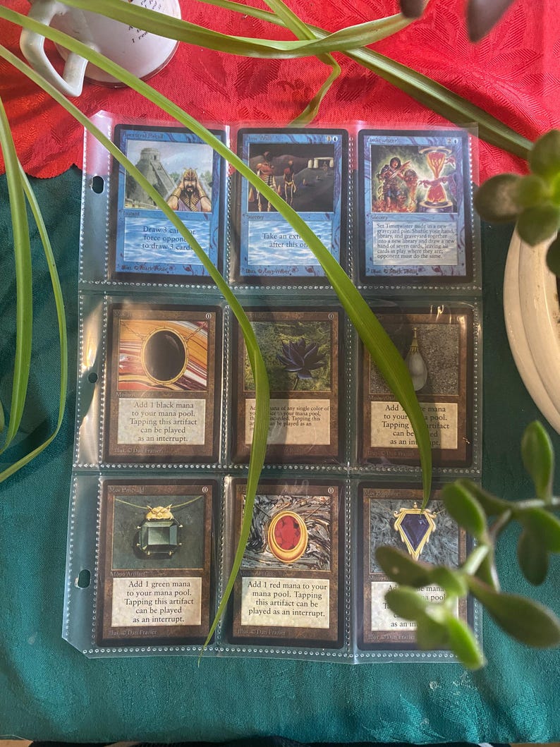 May include: A collection of Magic: The Gathering cards in a binder. The cards are all black bordered and feature various illustrations of artifacts and spells. The cards are organized by color and type.