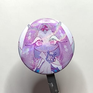 K&ouml;nnte beinhalten: Ein runder Ansteckbutton mit einem Anime-Charakter in Lila- und Rosat&ouml;nen. Der Charakter tr&auml;gt eine Krone und h&auml;lt eine Harfe, mit dem Text "Utger". Der Button hat einen gl&auml;nzenden, holografischen Effekt.