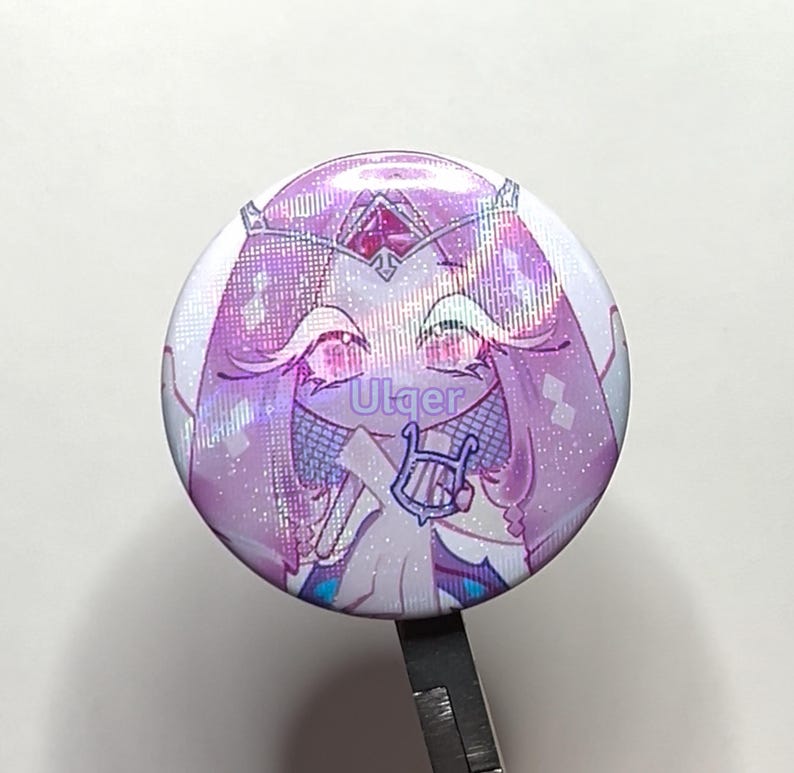 K&ouml;nnte beinhalten: Ein runder Ansteckbutton mit einem Anime-Charakter in Lila- und Rosat&ouml;nen. Der Charakter tr&auml;gt eine Krone und h&auml;lt eine Harfe, mit dem Text "Utger". Der Button hat einen gl&auml;nzenden, holografischen Effekt.