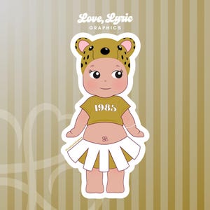 May include: A sticker featuring a cartoon baby wearing a gold crop top with "1985" printed on it, a white skirt, and a leopard print hat. The sticker has the text "Love, Lyric GRAPHICS" in the top left corner.