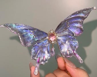 May include: A shimmering butterfly brooch with iridescent wings in shades of blue and purple. The center features a pink gemstone surrounded by gold filigree. A delicate gold pin extends from the center, and a small pink crystal hangs below.
