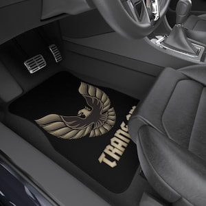 May include: Black car floor mat featuring a gold and black phoenix design with the text "TRANS AM". The mat is rectangular and sits in the driver's side footwell of a car.