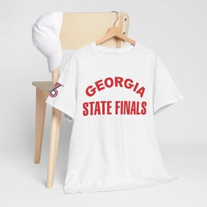 Pode incluir: Uma camiseta branca com o texto vermelho "GEORGIA STATE FINALS" em arco. Um boné branco está em uma cadeira de madeira. A camisa tem um pequeno logotipo na manga esquerda.