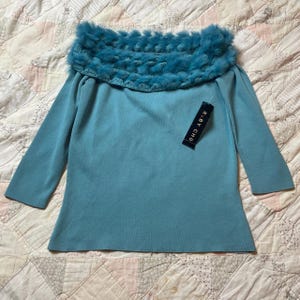 May include: A teal sweater with a faux fur trim around the neckline. The top has three-quarter sleeves and a tag that reads "RUBY CHO". The garment is made of a soft material.